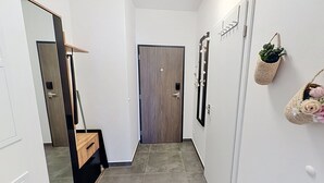 Un couloir étroit avec une porte en bois, un miroir, une plante fixée au mur et un petit panier rempli de fleurs.