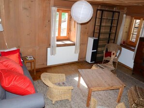 Ein Holzinnenraum mit einer Couch, zwei Korbstühlen, einem kleinen Tisch, einem Fernseher und einem Fenster mit Vorhängen.
