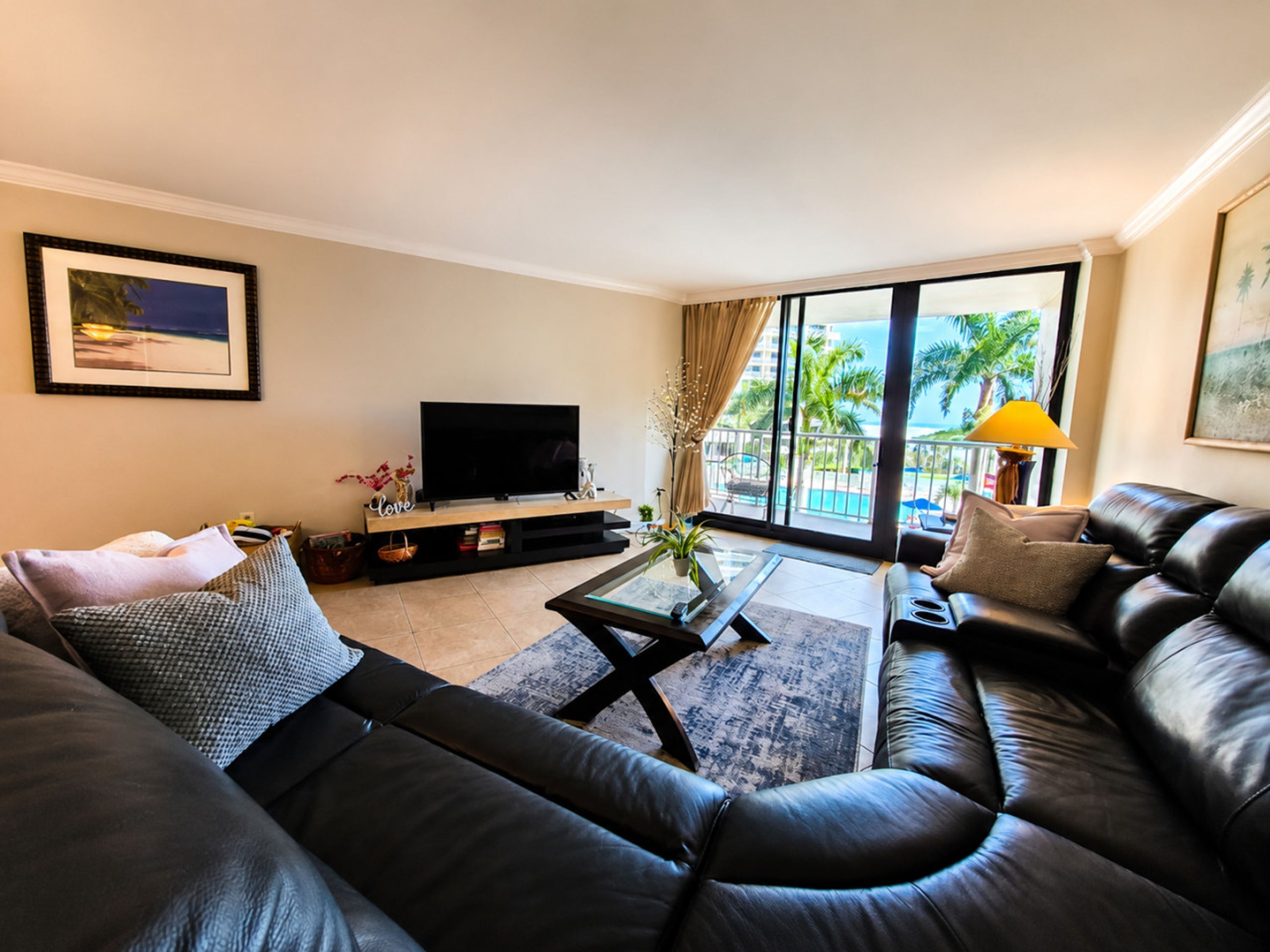 Ein Wohnzimmer mit einer schwarzen Ledersofa, einem Flachbildfernseher auf einem Holzboden, einem Couchtisch und einem großen Fenster mit Blick auf Palmen und einen Pool.