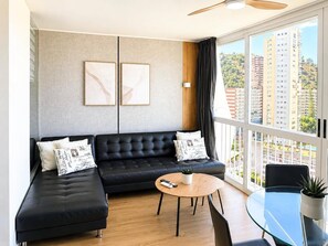 Ein modernes Wohnzimmer mit einer schwarzen Ledersofa, einem runden Holztisch und einem Glastisch für Essbereich. An der Wand hängen zwei gerahmte Kunstwerke, eine Deckenleuchte und ein Blick auf eine Stadtlandschaft durch große Fenster.