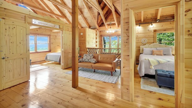 A wooden cabin interior with a bed, sofa, and a view of the outdoors. - Blue Mt. Ridge Cabin with Long Range Views, Hot Tub & Fire Pit Fun near Parkway! (Watauga County)