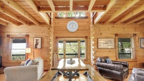 A wooden cabin interior with a wooden table, two sofas, a clock on the wall, and large windows. - Blue Mt. Ridge Cabin with Long Range Views, Hot Tub & Fire Pit Fun near Parkway! (Watauga County)