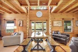 A wooden cabin interior with a wooden table, two sofas, a clock on the wall, and large windows. - Blue Mt. Ridge Cabin with Long Range Views, Hot Tub & Fire Pit Fun near Parkway! (Watauga County)