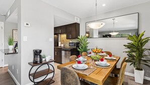 A modern dining area with a wooden table, chairs, and a kitchen in the background. - The Stirrup | New | Near Old Town + Pools (Scottsdale)