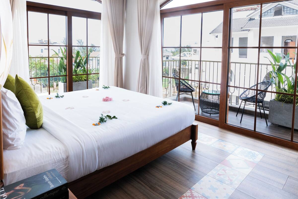 Habitación de hotel con una cama grande, ventilador de techo, balcón con sillas y vistas al exterior.
