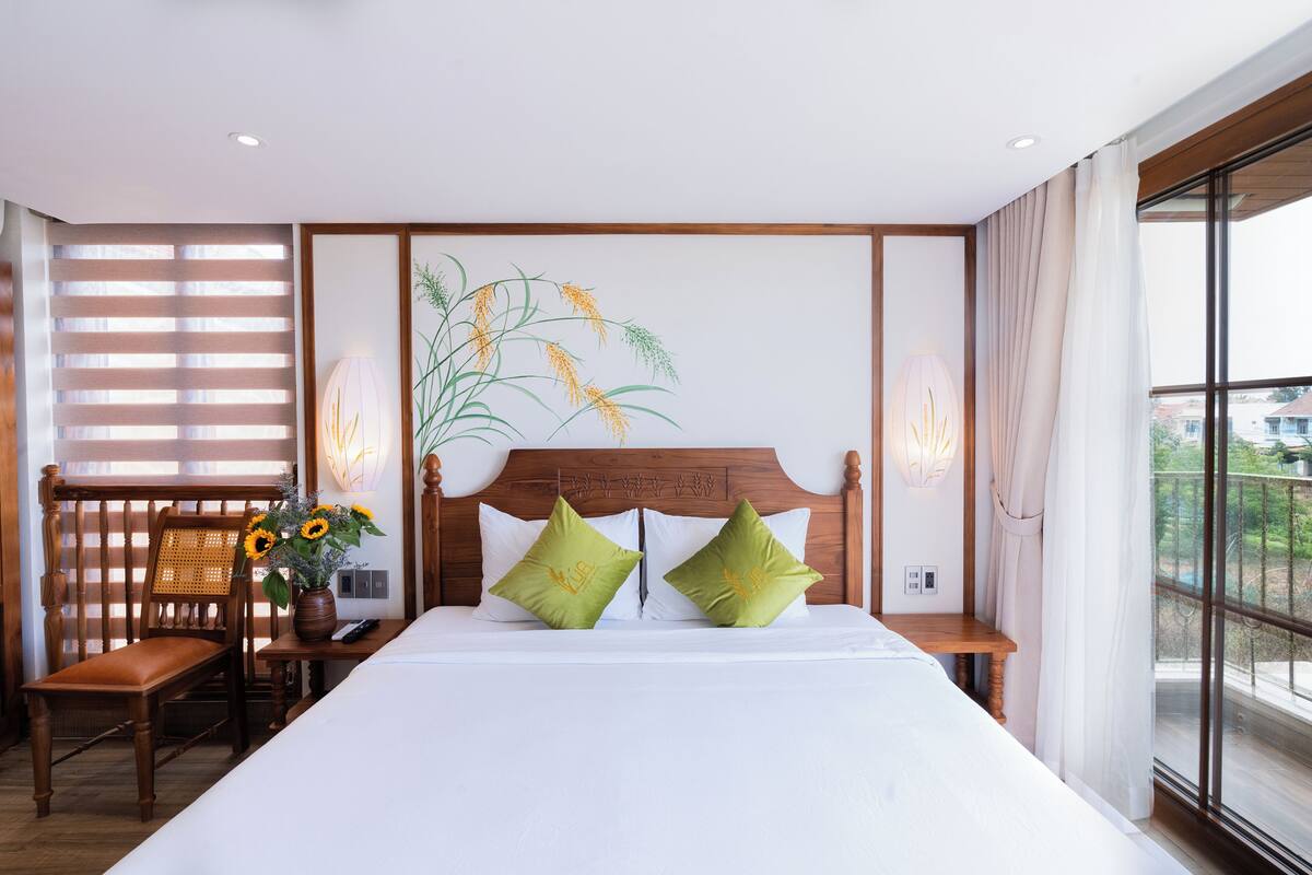 Habitación de hotel con una cama grande, dos almohadas verdes, un cabecero de madera, una mesita de noche, una silla, un jarrón con flores y una ventana con vistas.