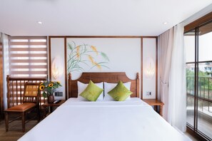 Habitación de hotel con una cama grande, dos almohadas verdes, un cabecero de madera, una mesita de noche, una silla, un jarrón con flores y una ventana con vistas.