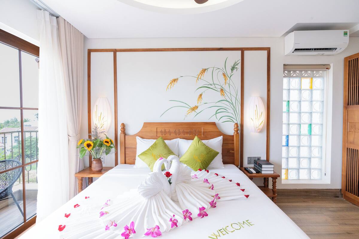 Una habitación de hotel con cama, mesita de noche, lámpara, un florero con flores, ventana con vistas y un equipo de aire acondicionado.