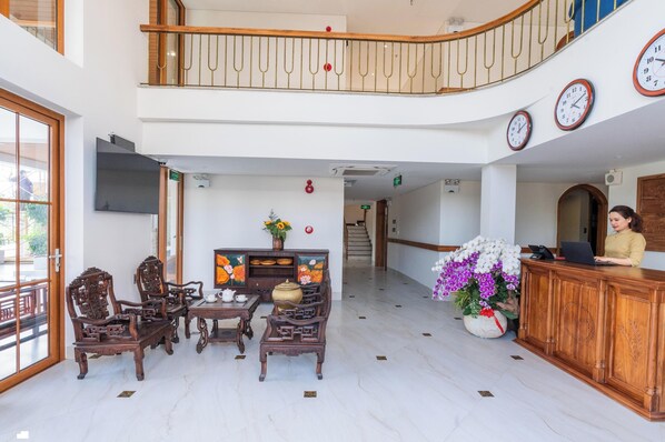 Vestíbulo de hotel con mostrador de recepción, sillas de madera, una mesa con tetera, un jarrón con flores y una escalera que conduce a un nivel superior.