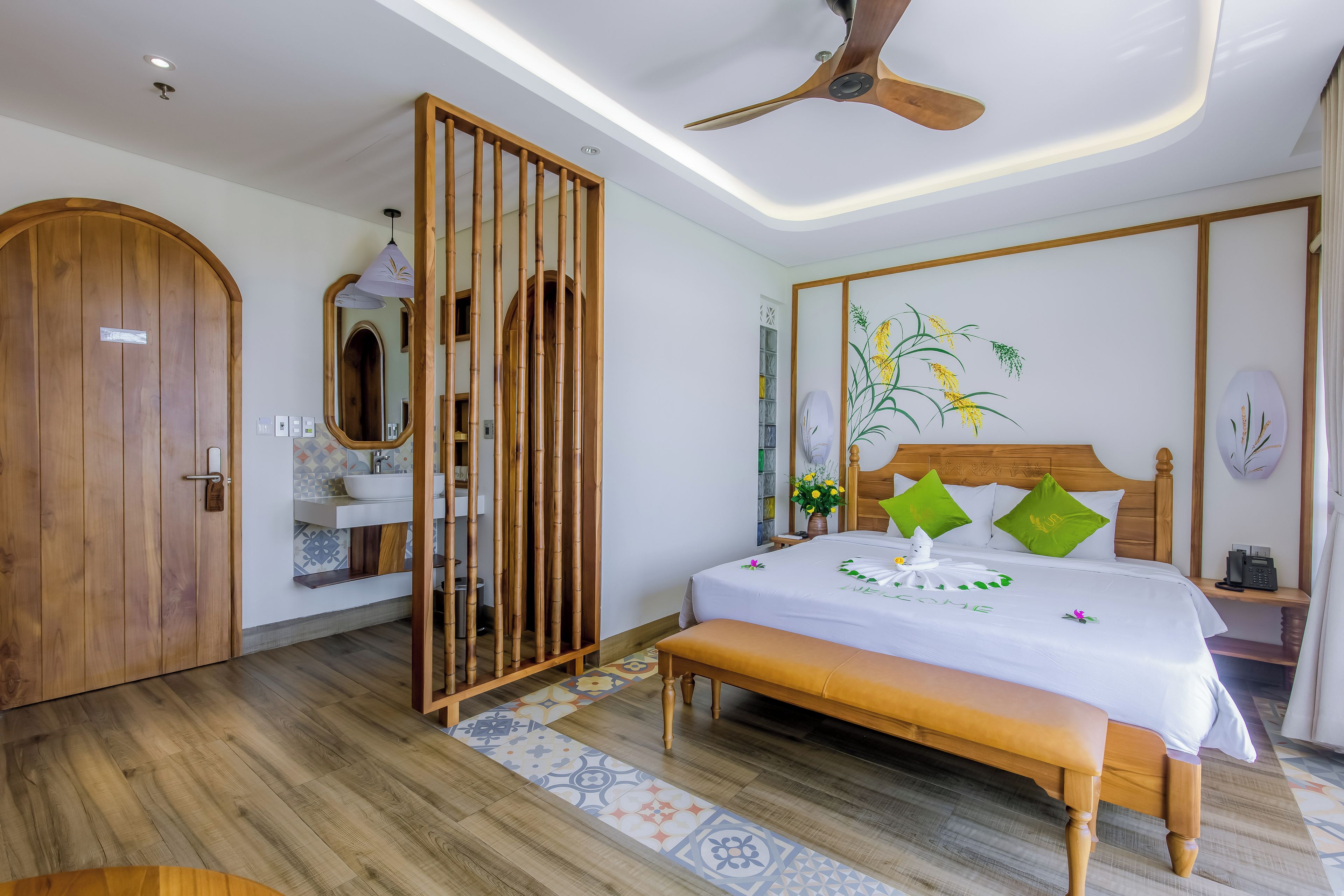 Quarto de hotel com porta de madeira, cama grande com colcha branca, ventoinha no teto e telefone fixado na parede.