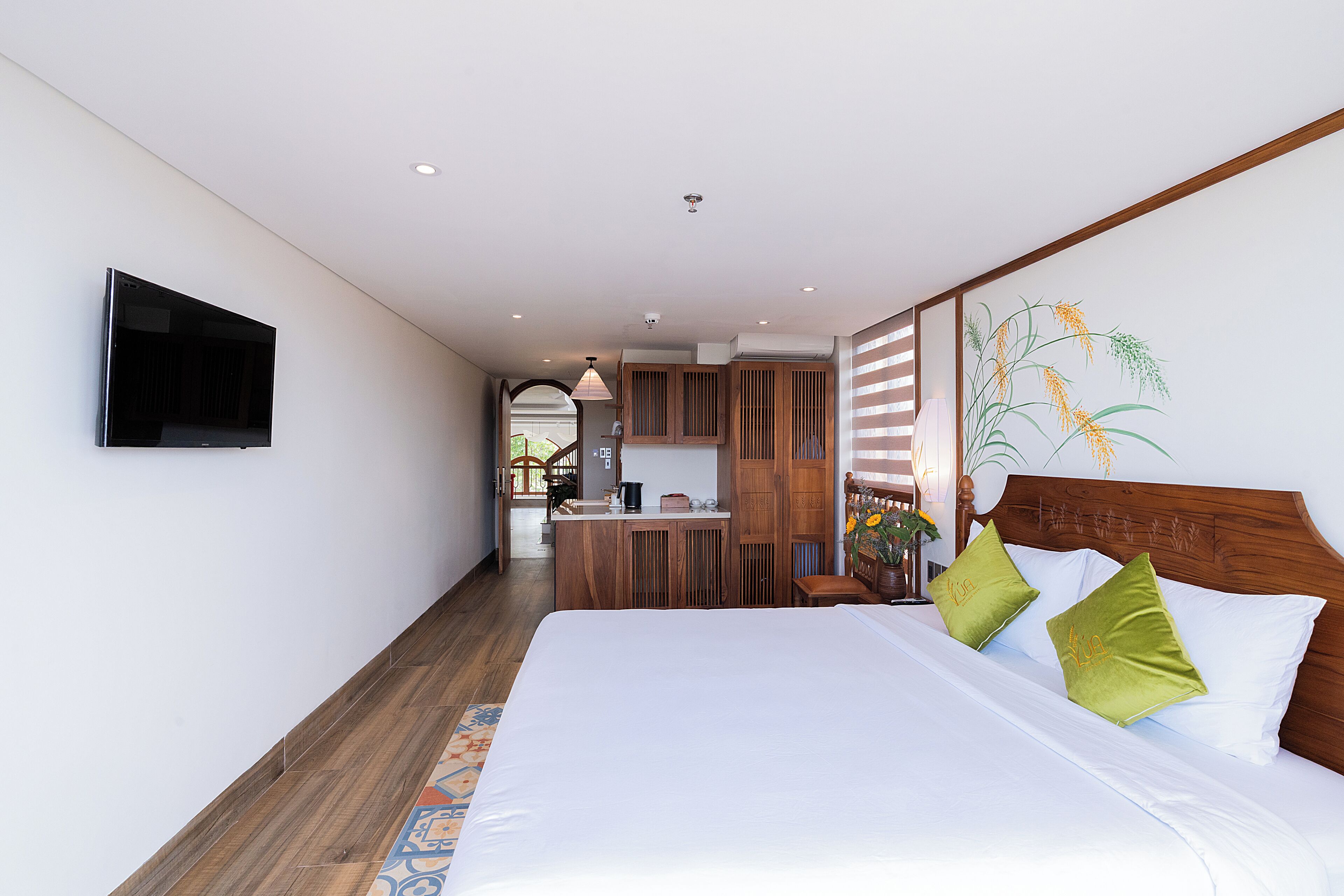 A bedroom with a bed, a TV mounted on the wall, and a kitchen area in the background.