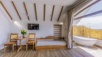 A bedroom with a bed, two chairs, a table with a vase of flowers, a TV, and a large window with a view.