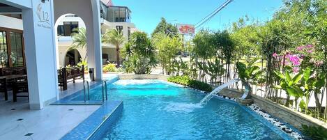 A swimming pool with a tiled edge, surrounded by greenery and a building with a balcony.