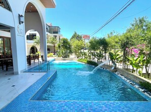 Una piscina con borde de azulejos, rodeada de vegetación y un edificio con balcón.