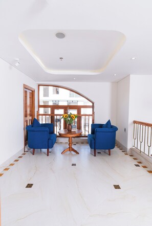 A spacious room with a high ceiling, a wooden door, two blue armchairs, a small table with a vase, and a view of a building through a large window. - Lua Boutique Hotel & Spa - An Bang Beach (Da Nang)