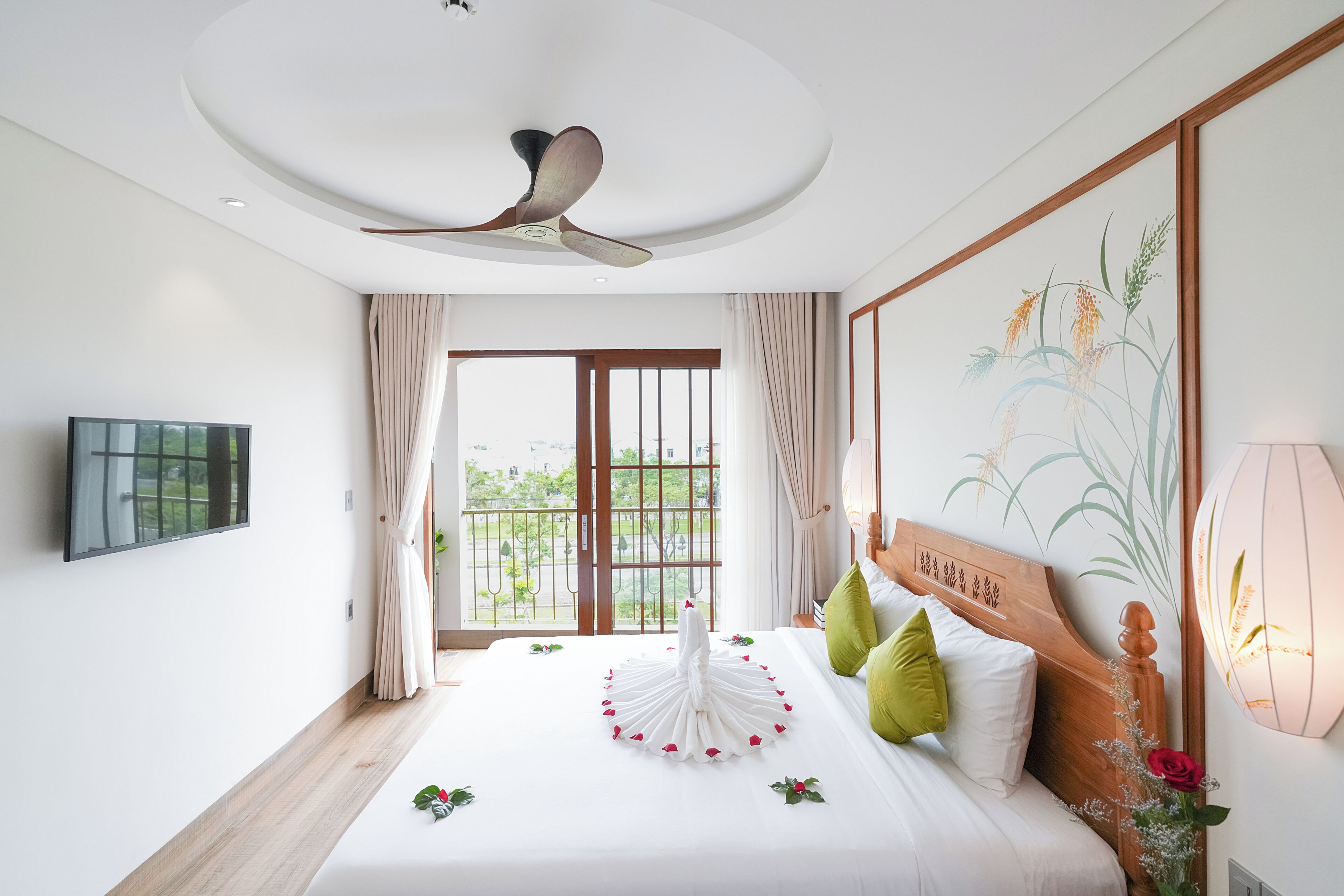 A hotel room with a bed, a ceiling fan, a TV, a window with curtains, and a wall-mounted light fixture.