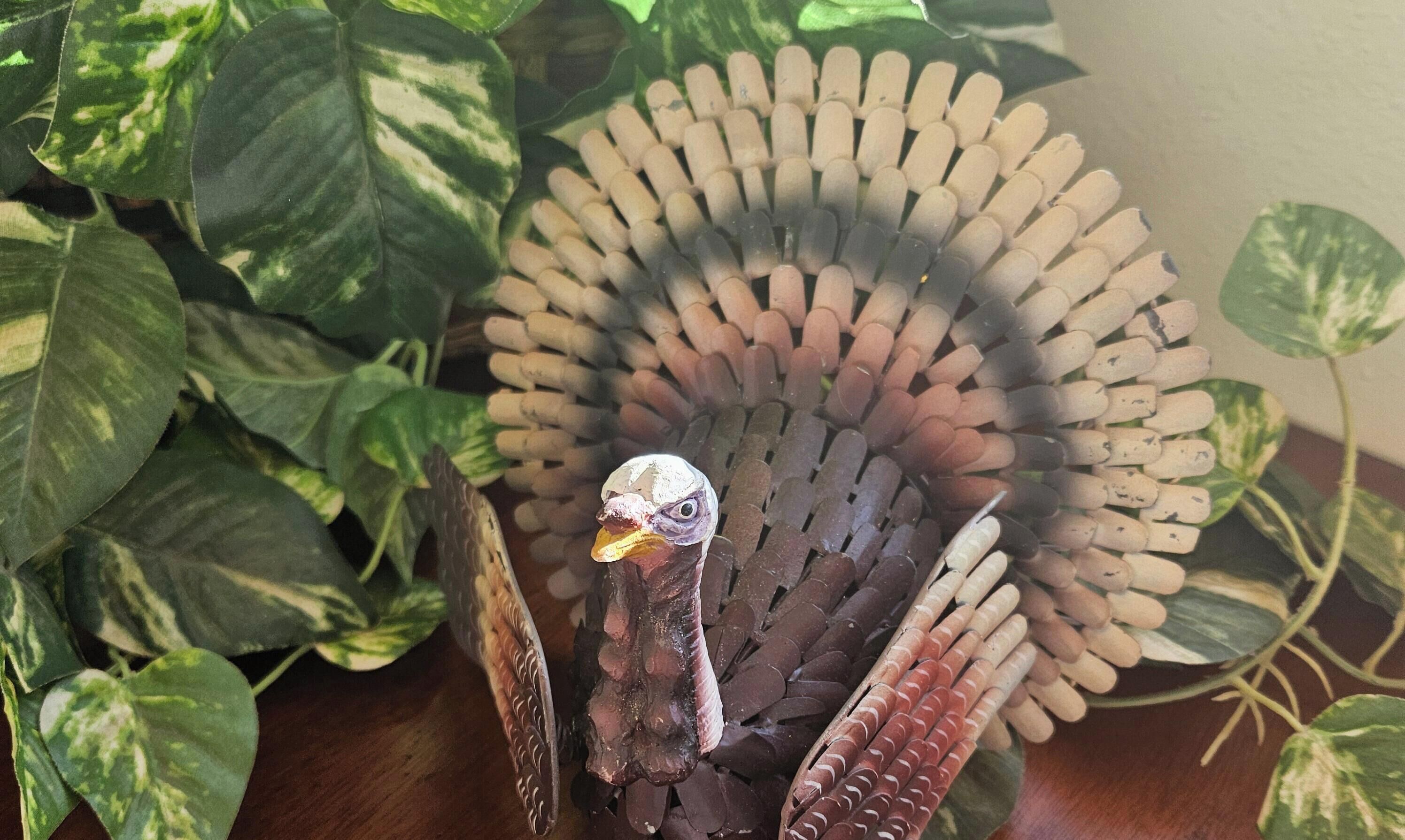 Una figurita de pavo con un abanico de palitos de madera, rodeada de plantas de hojas verdes.