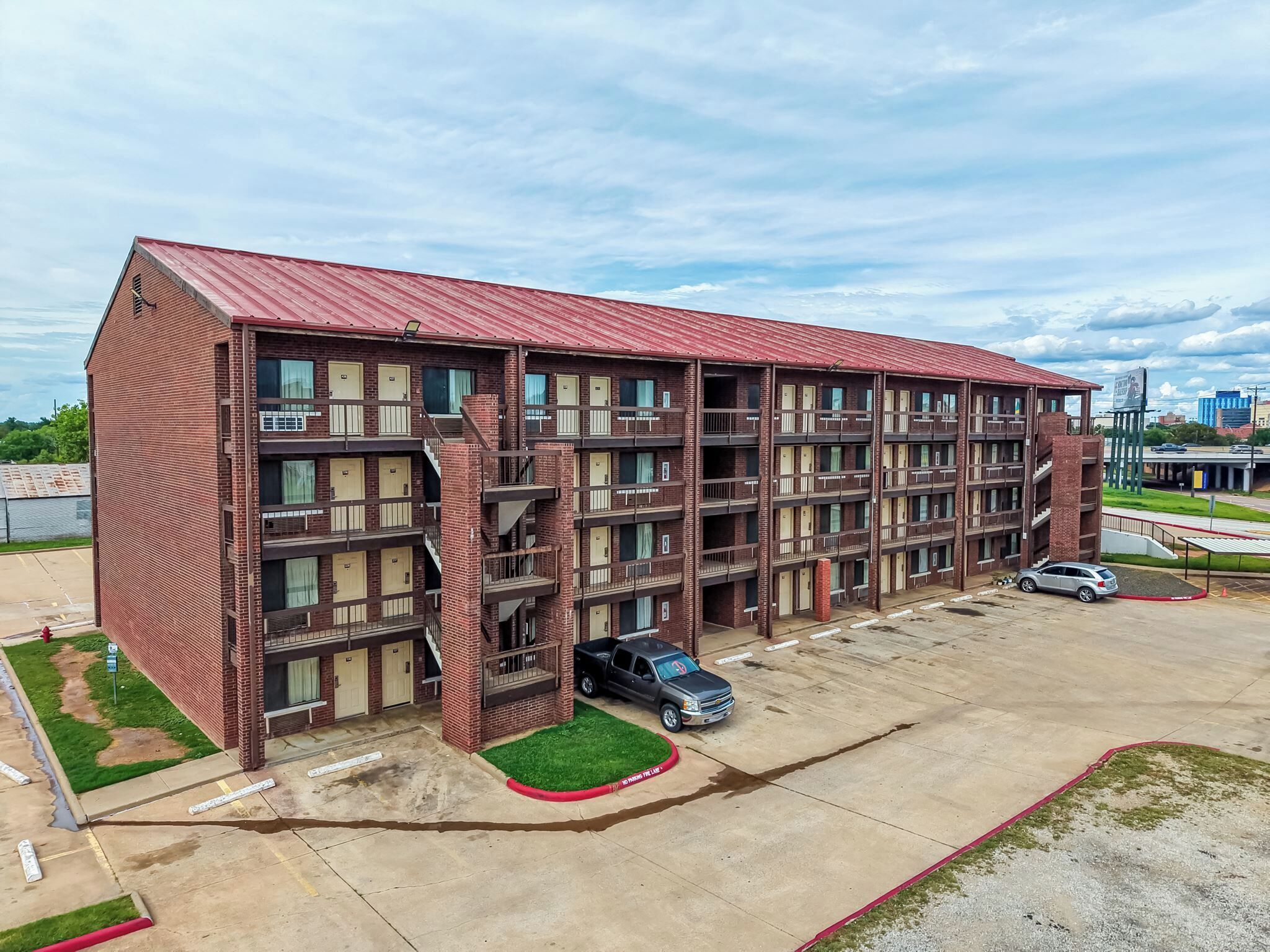 Un edificio in mattoni a più piani con tetto rosso, diversi balconi e un parcheggio.