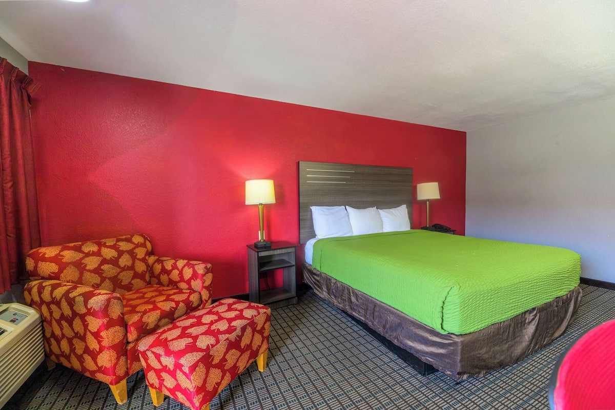 Habitación de hotel con una pared roja, una cama con colcha verde, dos sillas con estampado de hojas y dos lámparas de noche.