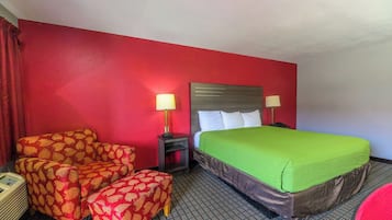 Habitación de hotel con una pared roja, una cama con colcha verde, dos sillas con estampado de hojas y dos lámparas de noche.