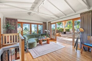 A spacious living area with a green sofa, wooden flooring, and large windows offering a view of the outdoors.