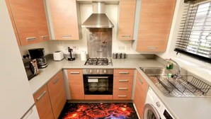 A compact kitchen with wooden cabinets, a stainless steel range hood, a built-in oven, a washing machine, and a window with blinds. - Stylish 2BR Apartment Parking PS5 Wifi (Darwen)