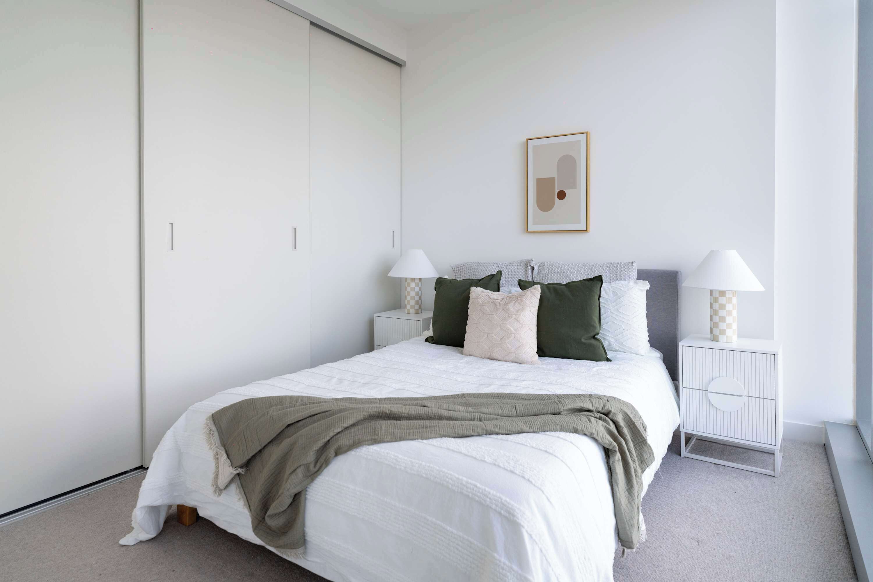 Une chambre à coucher comprenant un lit, deux tables de chevet avec des lampes, une œuvre d’art accrochée au mur et un placard à portes coulissantes.