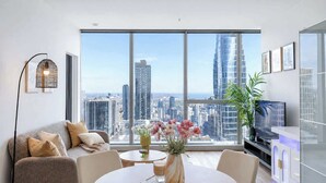 A modern living room with a city view, a sofa, a coffee table, a vase with flowers, and a TV. - Sky High Views CBD Stay Near Ritz Carlton (Melbourne)