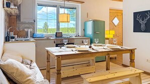 A cozy kitchen with a wooden ceiling, a dining table set with plates and glasses, a blue refrigerator, and a window with a view of greenery.