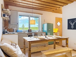 A cozy kitchen with a wooden ceiling, a dining table set with plates and glasses, a blue refrigerator, and a window with a view of greenery.