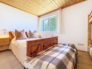 Un dormitorio con techo de madera, una cama grande con manta a cuadros, una mesita de noche con lámpara y una ventana con cortinas.