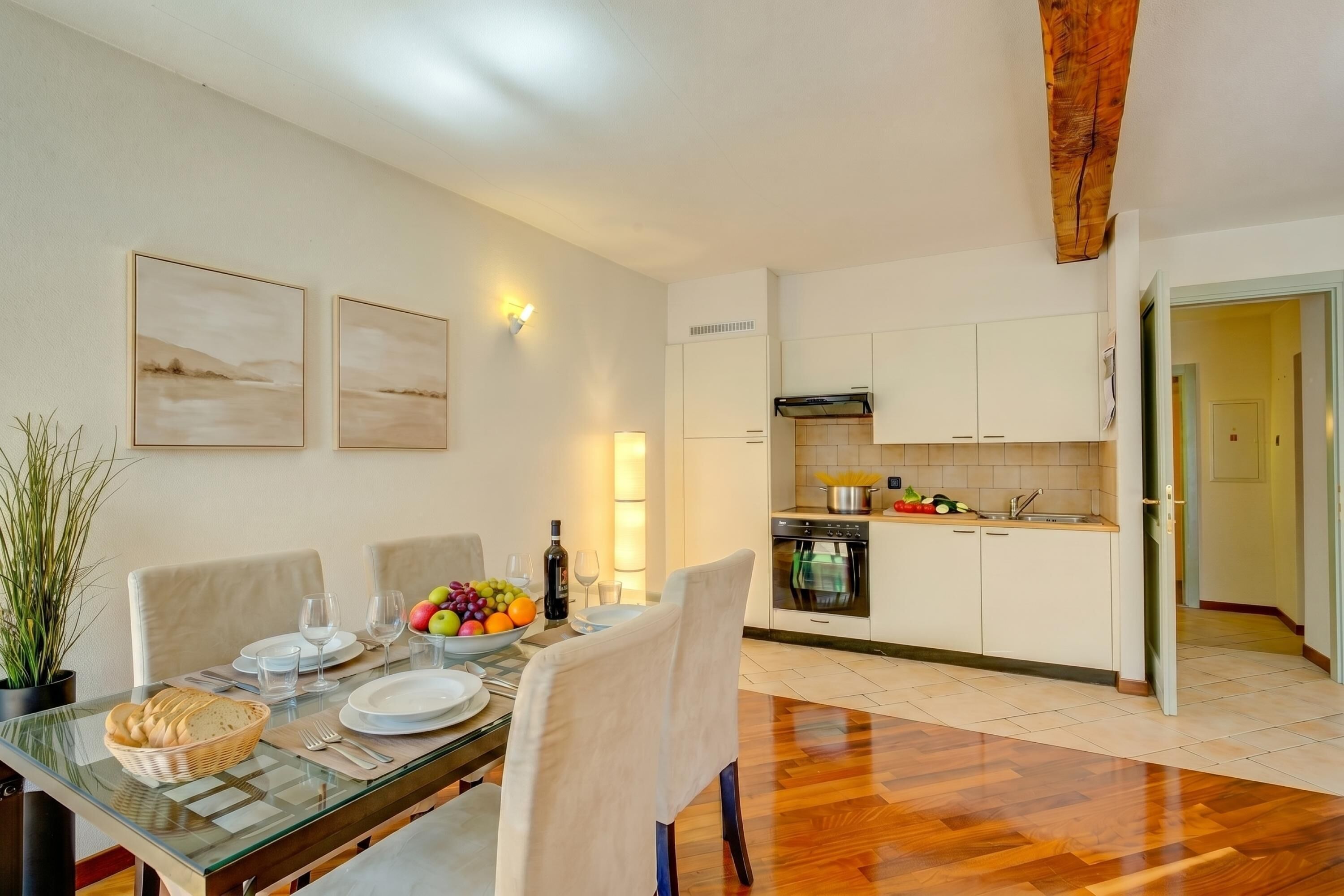 A dining area with a glass table, chairs, and a fruit bowl. There is a kitchen with white cabinets and a stove.