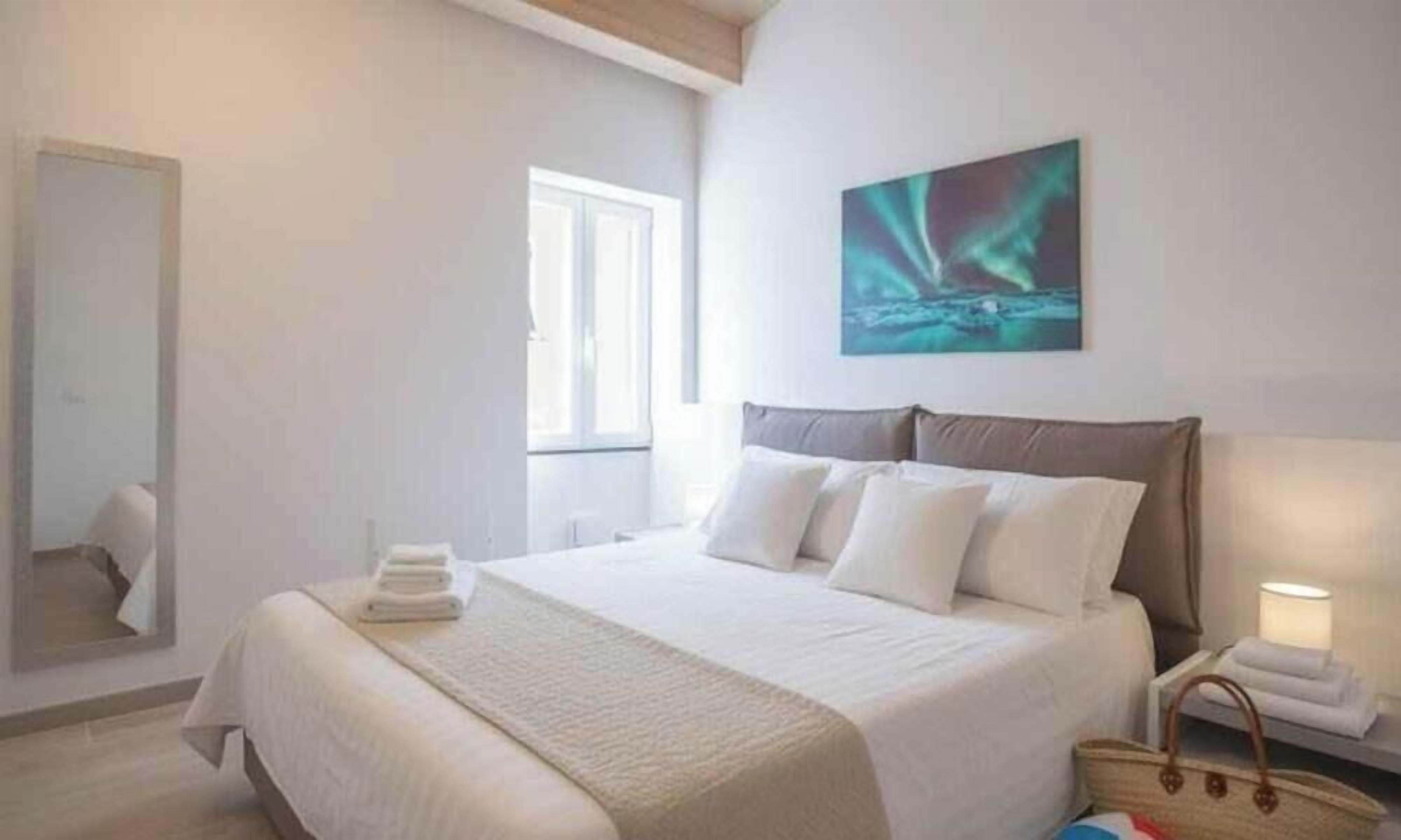 Ein Schlafzimmer mit Bett, Kissen, Nachttisch, Lampe, Spiegel und einem Bild an der Wand.