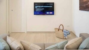 Un salon avec un téléviseur à écran plat fixé au mur, un panier en osier contenant un tissu à rayures et un canapé beige agrémenté de plusieurs coussins.