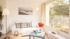 Ein Zimmer mit einem Bett, einem kleinen Tisch mit Vase und Büchern, einer Lampe und Blick auf einen Garten durch ein Fenster.