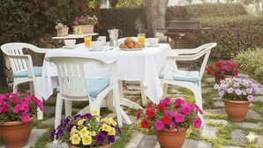 Ein Essbereich im Freien mit einem für das Frühstück gedeckten Tisch, umgeben von weißen Stühlen und Kübelblumen.