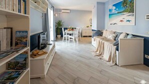Un salon comprenant un canapé, une table à manger avec des chaises, une bibliothèque et un tableau représentant une scène de plage.