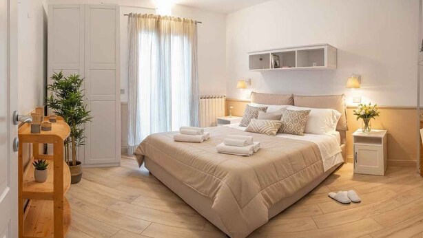 Une chambre à coucher avec un lit, des tables de chevet, une commode, une plante en pot, une étagère en bois et une fenêtre avec des rideaux.