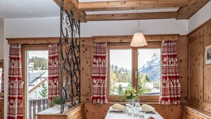 A wooden interior with a table set for dining, large windows with curtains, and a view of mountains. - Chesa Minchületta by Interhome (Silvaplana-Surlej)