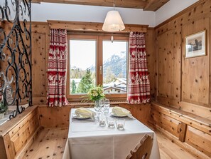 A wooden interior with a dining table set for two, a window with curtains, and a framed picture on the wall.