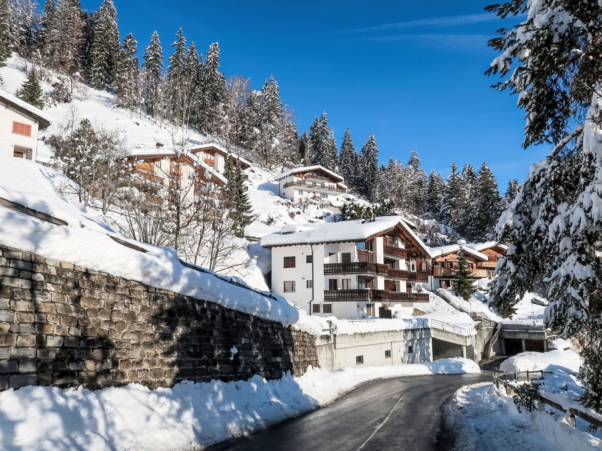A snow-covered mountain village with chalet-style buildings, a stone wall, and a clear blue sky.