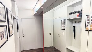 Un pasillo moderno con suelo de madera, paredes blancas y un panel de vidrio. Hay fotografías enmarcadas en las paredes y un perchero con un paraguas.