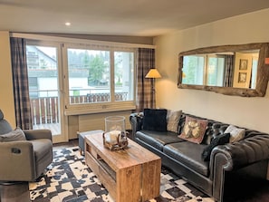 A living room with a sofa, a wooden coffee table, a lamp, and a large window with curtains. - Val Signina 6-7 by Interhome (Laax)
