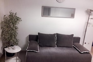 Ein modernes Schlafzimmer mit einem grauen Sofa, zwei Kissen, einem kleinen Beistelltisch, einer Topfpflanze und einem Spiegel an der Wand.