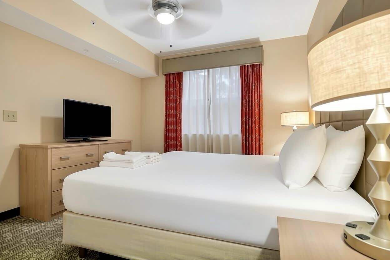 Quarto de hotel com cama, televisão, luminária e janela com cortinas.