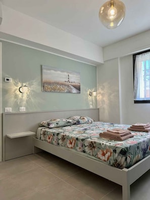 Ein Schlafzimmer mit einem Bett, einer Wandleuchte, einem Gemälde und einem Fenster mit Vorhängen.