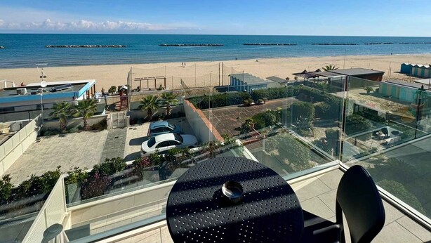 Um edifício alto com vista para a praia, uma área de estacionamento e um espaço de refeições com mesa e cadeiras.
