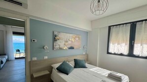 Quarto de hotel com cama, pintura na parede, janela com cortinas e vista para o mar através de uma porta aberta.