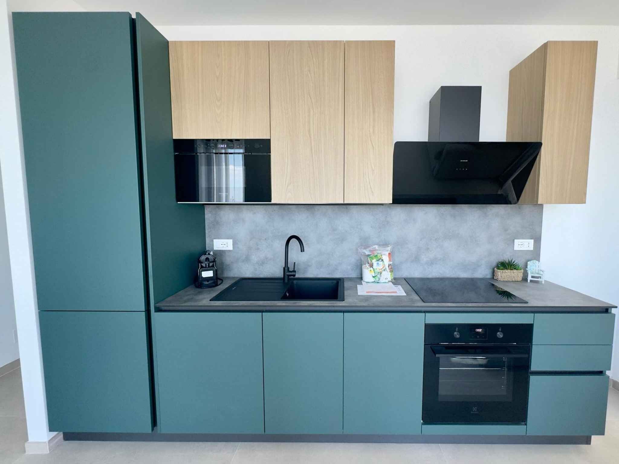 A modern kitchen with teal cabinetry, a black sink, and a built-in oven.