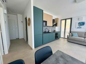 A modern kitchen with teal cabinets, a dining area with a table and chairs, and a living room with a sofa and a painting. - Holiday apartment with air conditioning, internet and balcony with sea view (Pesaro)